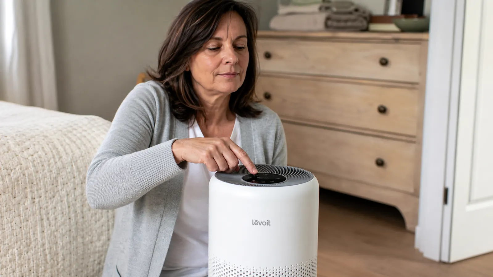 Hand adjusting the speed dial on top of a white Levoit air purifier on a bedroom dresser