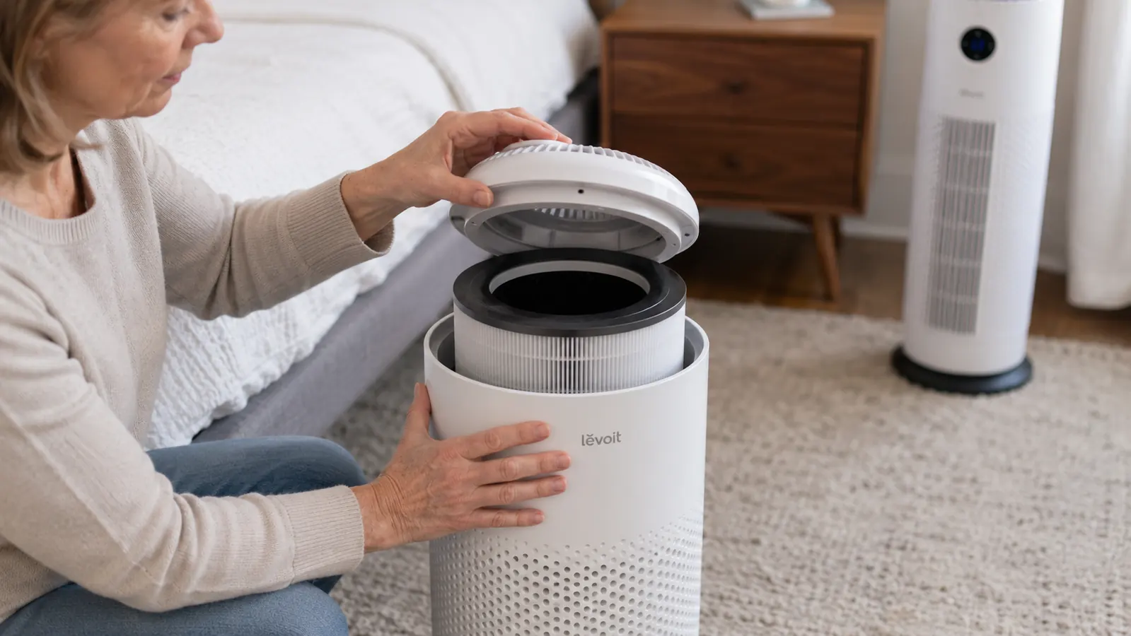 Hand lifting the top panel off the Levoit air purifier to show the cylindrical filter underneath, with a tower style purifier blurred in the background