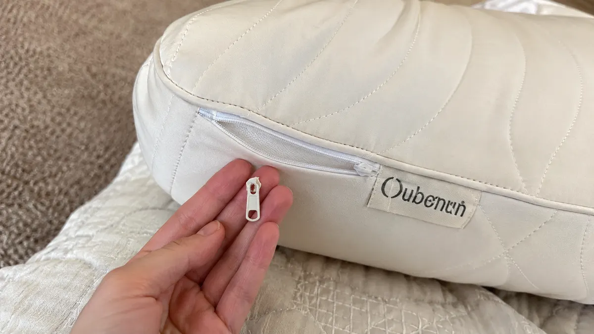 Close-up of a hand holding a small broken zipper pull tab beside the corner seam of a quilted body pillow